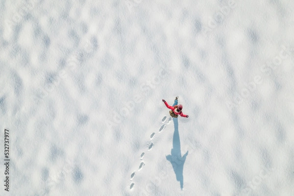 Fototapeta Woman in a red christmas jacket with backpack walking on the snow and doing footprints in a park. Winter. Drone, top, aerial view.
