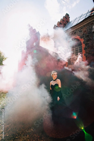 Fototapeta Art work. A slender beautiful woman with blond hair in a black vintage dress and expensive jewelry, stylish makeup and hairstyle, comes out of an ancient castle in fire and smoke.