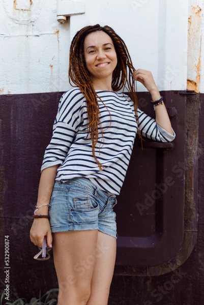 Obraz a girl with a dreadlocked hairstyle poses in the summer outdoors, dressed in a T-shirt and denim shorts