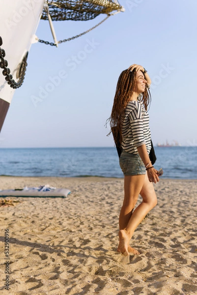 Obraz a girl with a dreadlocked hairstyle poses on the beach near a ship in summer, the sea and sand on a bright sunny day, dressed in a T-shirt and denim shorts