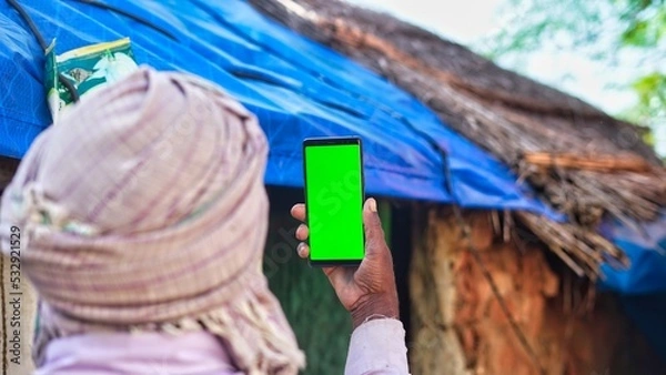 Fototapeta Old Indian man taking selfie and showing a green screen cell phone with rural village life concept background.