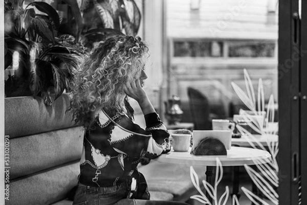 Fototapeta Beautiful Woman Sitting at Table in Cafe. Pretty Young Girl With Curly Hair Looks Out Window While Sitting In Coffee House