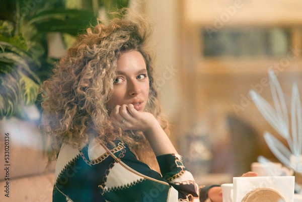 Fototapeta Portrait of Beautiful Woman Sitting at Table in Cafe. Pretty Young Girl With Curly Hair Looks Out Window While Sitting In Coffee House