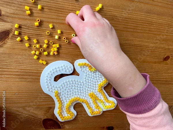 Obraz Hand eines kleinen Kinde, das gelbe Steckperlen auf eine Katzenform aufsteckt an einem Holztisch zur Beschäftigung