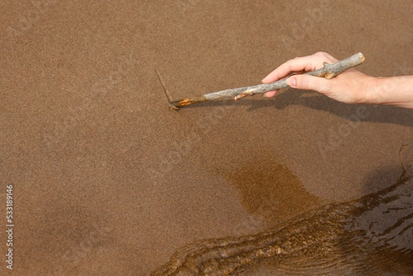 Obraz Middle-aged woman writing on the wet sand on the beach near water. Human hand with wooden stick scratching a line