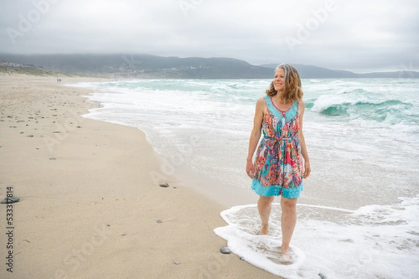 Obraz An adult woman walks along an empty and wild beach with waves from the sea.