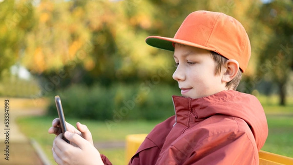 Fototapeta A teenage boy sits on a wooden bench in the park in autumn and uses a mobile phone, watches videos, surfs the Internet. Video games, social media addiction.