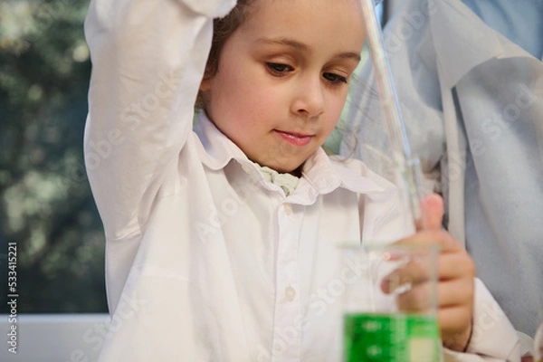 Obraz Beautiful mischievous child girl holding a glass pipette, and dripping few drops of green chemical solution from a flask into a test tube during a chemistry lesson, in the school science laboratory