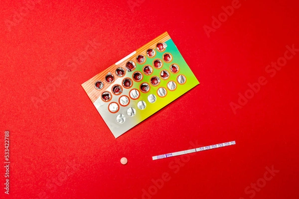 Fototapeta A purple and white single-strip pregnancy test and birth control pills lie on a red background. Menstrual cycle and pregnancy. Negative pregnancy test and contraceptives