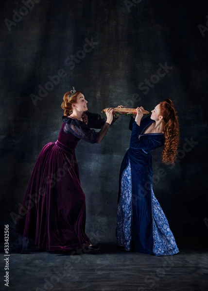 Fototapeta Portrait of two beautiful women in image of queens fighting for giant sandwich isolated over dark background