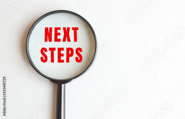 Fototapeta NEXT STEPS text written on gray background. We read it through a magnifying glass.