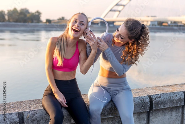 Fototapeta Two woman friends with headpohones taking break between jogging and listen music and have fun