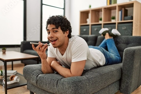 Fototapeta Young hispanic man talking on the smartphone lying on the sofa at home.