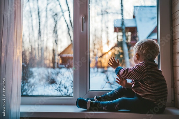 Fototapeta little boy looks out the window at home in winter waving his hand and feeling sad on a cold day