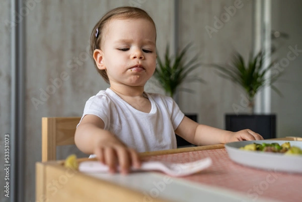 Fototapeta One baby girl small caucasian toddler eating while sit at the table alone at home copy space little child having lunch healthy meal in room in day taking fork