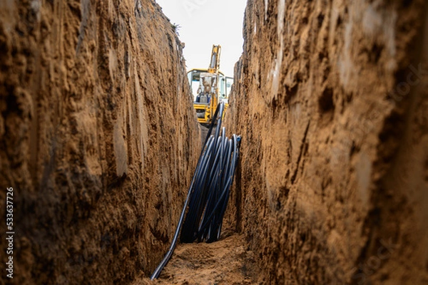 Obraz Installation of HDPE pipes in the trench. The process of laying of engineering systems water supply. Black plastic pipes underground.