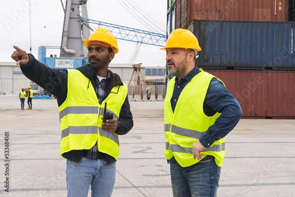 Fototapeta Two dock workers in reflective waistcoats and hard hats talking to each other outdoors