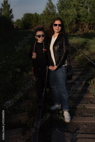 Obraz A young mother and son in sunglasses stand holding hands on the railroad tracks against the backdrop of the forest. a family of two