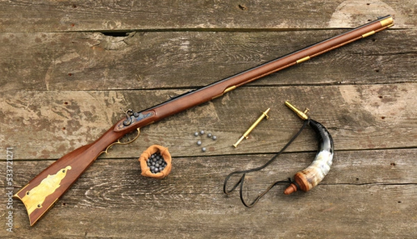 Obraz Traditional Kentucky rifle and shooting equipment such as a gunpowder horn, a gunpowder measure and bullets on an old, damaged board.
