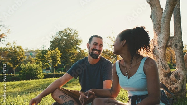 Fototapeta Happy smiling interracial couple talking while sitting on the grass in the park. Backlight