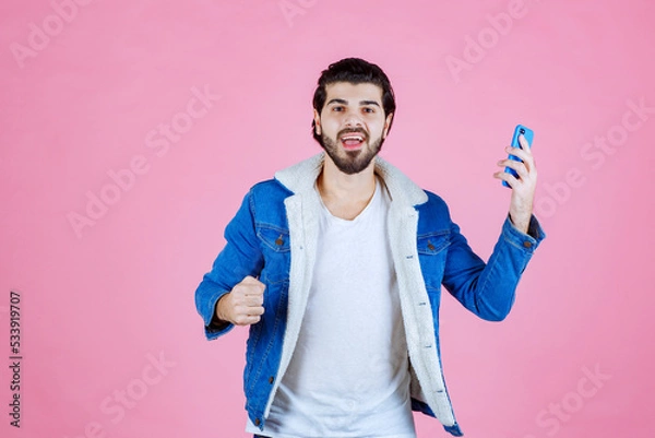 Obraz Young boy holding a smartphone and making a peace sign against a pink background, wearing a blue jacket and casual attire, showcasing confidence and modern technology usage