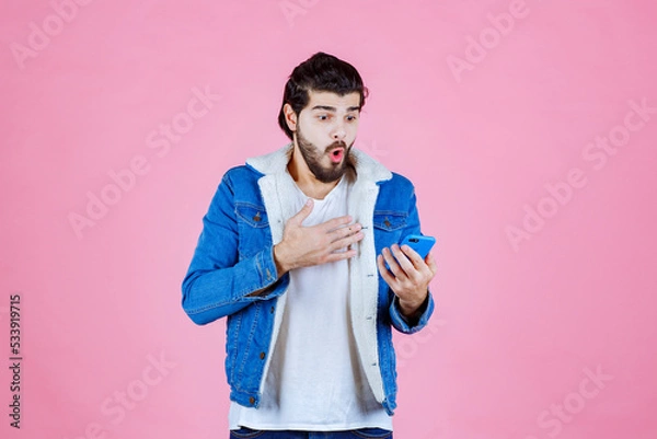 Obraz Young man using smartphone while standing against a pink background, casual fashion style, focused on technology and communication.