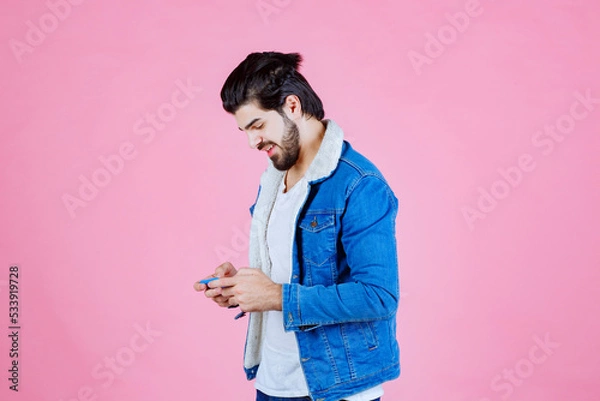 Obraz Young woman in a blue denim jacket holding a smartphone against a pink background, casual fashion, modern lifestyle, focused on technology use