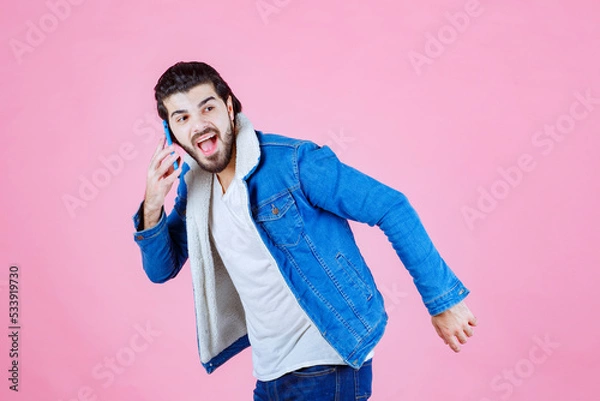 Obraz Young man in casual attire holding a smartphone against a pink background, expressing excitement or surprise with a dynamic pose and energetic stance.