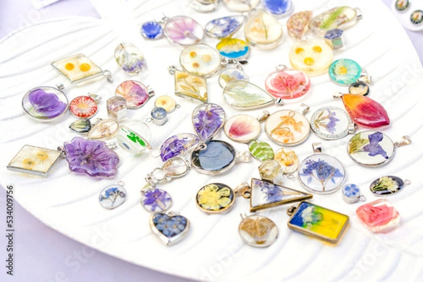 Obraz Epoxy resin jewelry - many handmade pendants with real flowers and plants inside on white stand. Making exclusive jewellery at home