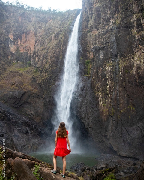Fototapeta girl in red dress stands under huge waterfall, dramatic view of long-haired woman in front o large waterfall wallaman falls, highest waterfall in australia