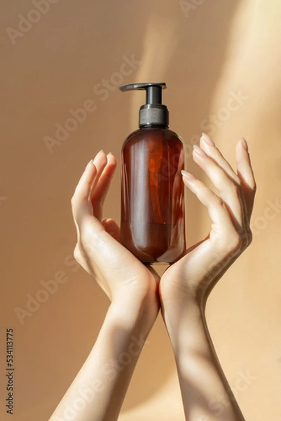 Obraz Bottle with dispenser, sprayer, for liquid soap, shampoo, lotion in women's hands on beige background. Rays of sunlight fall on container made of amber glass. Mockup concept, vertical frame
