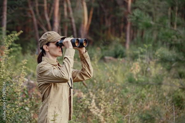 Fototapeta A woman park ranger in uniform looks through binoculars and monitoring the forest area in summer, selective focus.
