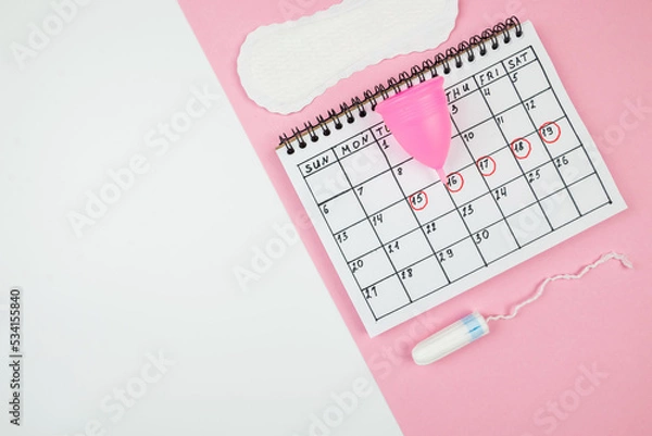 Fototapeta Top view of a calendar of women's critical days with pads and accessories on a white and pink background with copy space.