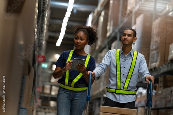 Obraz Young African America woman holding tablet to check the order while looking boxes and colleague pushing trolley to pick up shipment, multiethnic workers working in storage import-export warehouse