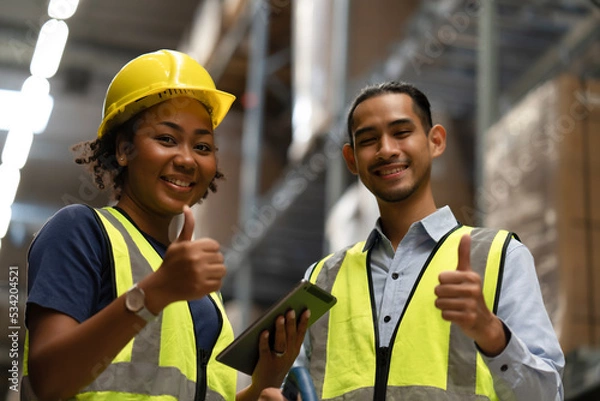 Fototapeta Black woman with yellow helmet and man warehouse worker show thumb up together for successful job with happy face. Team spirit working together in storehouse to complete the balance inventory