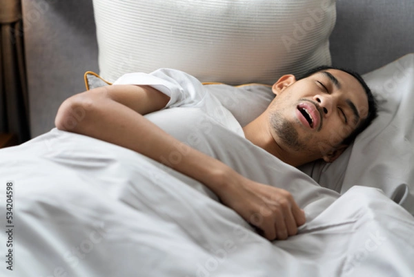 Fototapeta A young bearded man sleeps with his mouth open and makes a loud snore lying down on bed, tired and deep sleep. Making noise and annoying his partner.