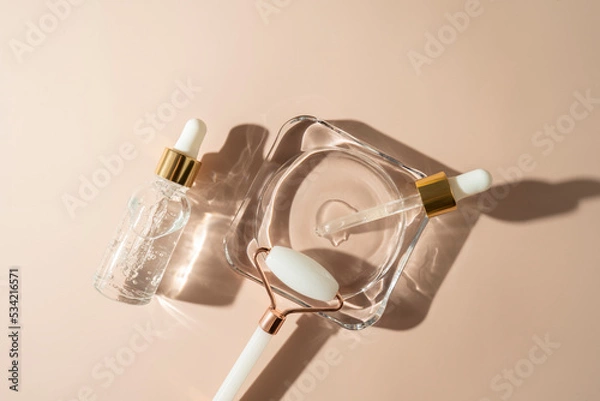 Fototapeta A transparent face serum in dropper bottle or essential oil and face roller lying on a glass plate on a beige background