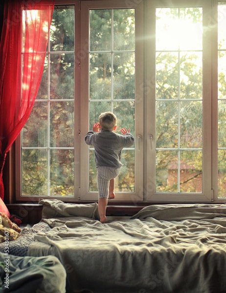 Fototapeta little boy looks out the window in a sunny room