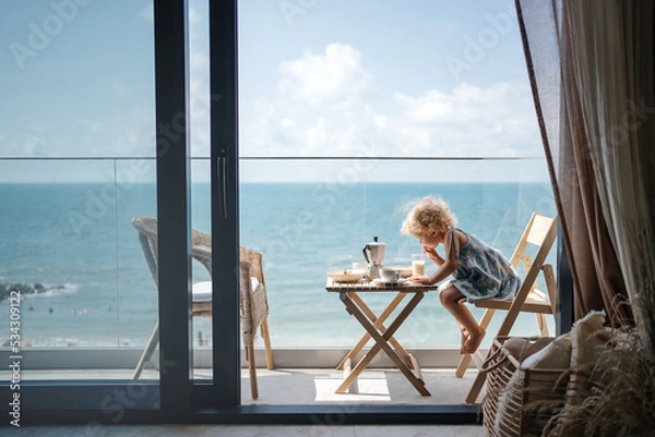 Fototapeta small child girl with blond curly hair has breakfast in the attic overlooking the seascape. Drinks milk.