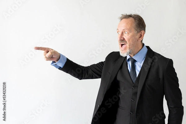 Obraz Portrait of angry mature businessman pointing at someone. Senior Caucasian manager wearing three piece suit looking away and shouting against white background. Angry boss concept