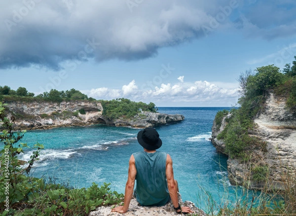 Obraz man sitting on the edge of a cliff with a view of a blue lagoon