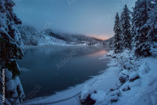 Fototapeta December winter sunrise in Tatra Mountains near lake. Thin ice covers the lake, fog go down the mountainside, fir trees covered snow. Houses are visible in the distance.