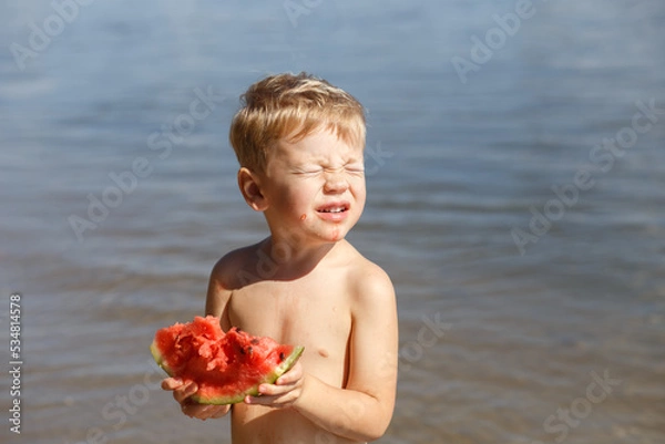 Fototapeta A blond boy 3-4 years old eats a piece of ripe red watermelon, looks at the bright sun, closes his eyes against the background of the river. On a sunny summer day