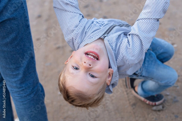 Fototapeta Cheerful blond boy 3-4 years old holds hands of adults and looks into the camera from the bottom up.