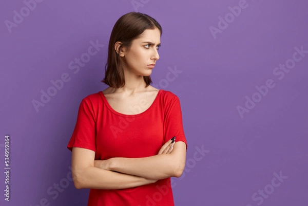 Fototapeta You are unfaithful. I dont believe you. Angry young pretty brunette girl in red t-shirt keeping her arms folded, looking aside with a stern and skeptical expression. Posing over purple background.