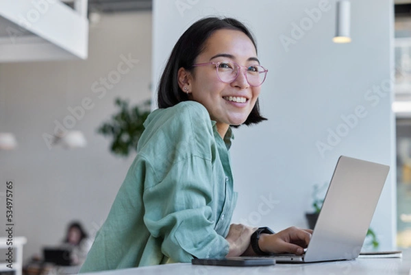 Fototapeta Pretty cheerful Asian woman in eyeglasses and casual clothes browses laptop computer connected to 4g internet updates software uses modern technologies poses in cafeteria looks gladfully away