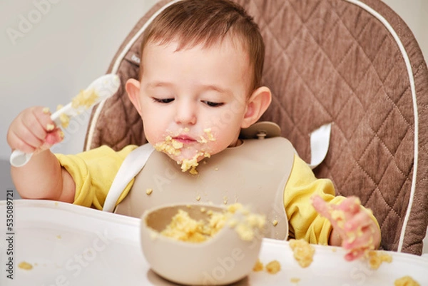 Fototapeta Happy toddler baby boy learns to eat porridge himself with a spoon while sitting in a child chair. Smiling child eats with his hands and a spoon from a plate, kid aged one year