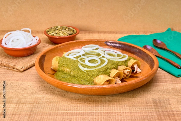 Obraz Mexican food (green mole enchiladas) served with onion rings in a clay dish on a wooden table, decorated with colorful napkins of the Mexican flag. copy space