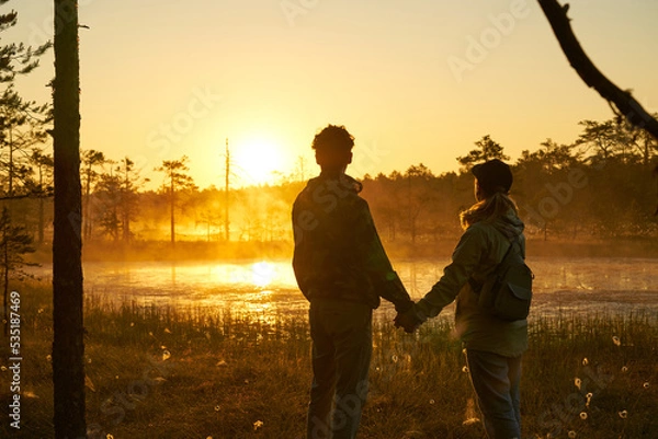 Obraz woman and man holds hands tourist meets dawn in nature. Sunset,  light and fog, Reflections of trees in lakes . Travel romance. Viru swamps Estonia.