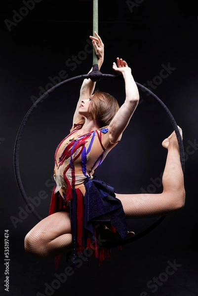 Obraz Beautiful woman performing acrobatic element on aerial ring indoors. Circus performer doing trick on arial lira on black background.